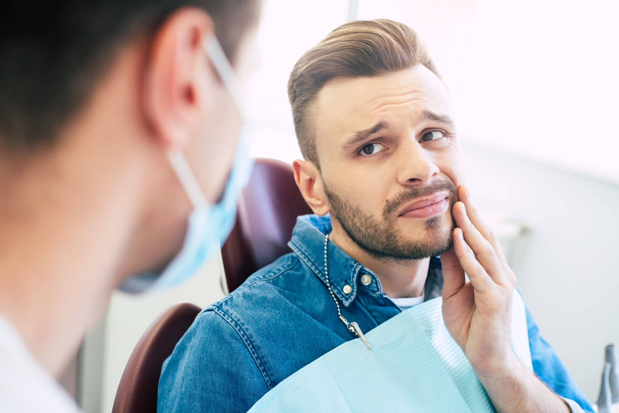 Symptoms of gums pain. A man with a worried face is holding his hand on his cheek because of irritating pain in front of a dentist who is going to give a patient a treatment. Man holding his jaw in pain due to impacted wisdom teeth, illustrating the need for wisdom teeth removal at Plainsboro Dental Services in Plainsboro, NJ.